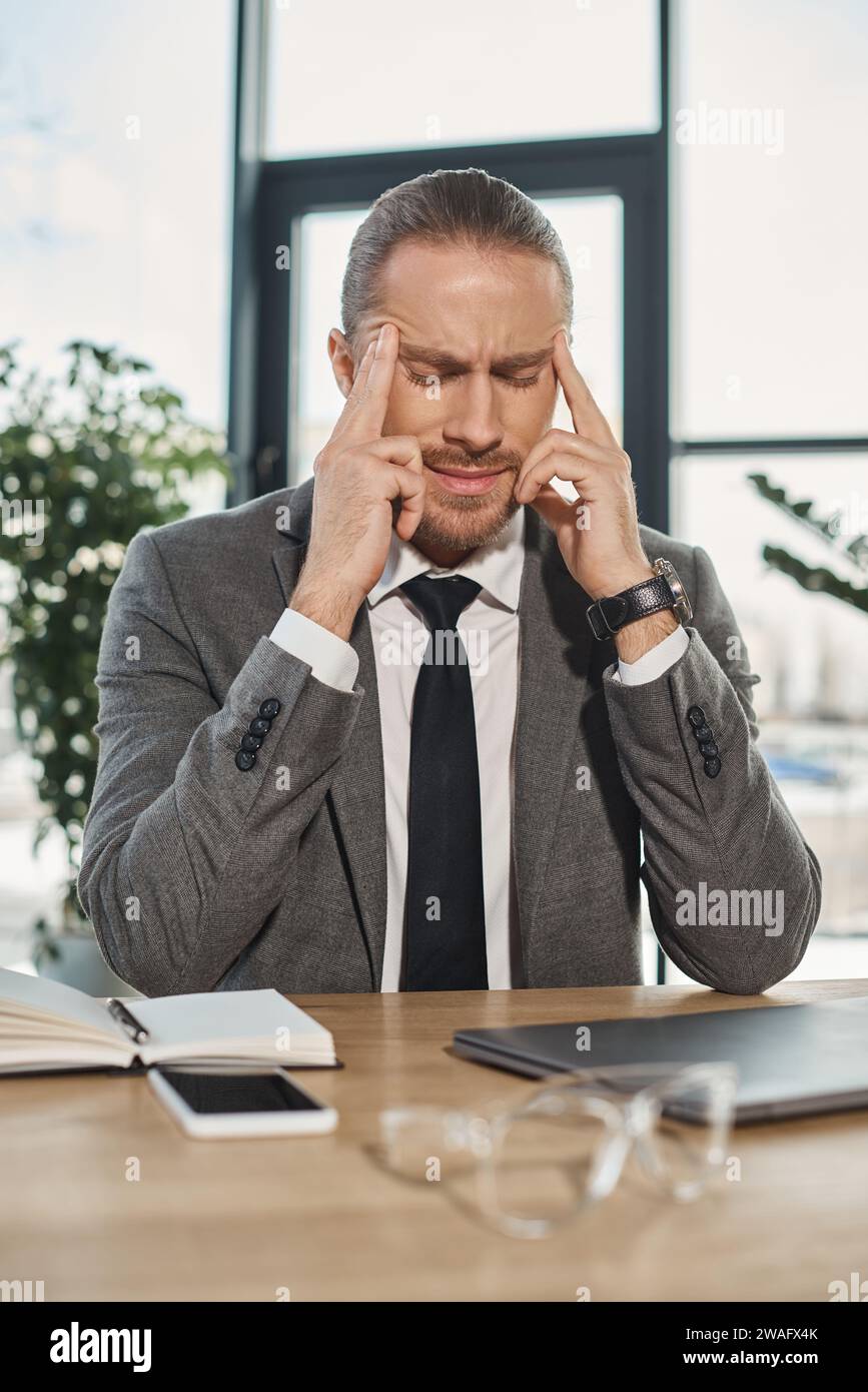 Müder Geschäftsmann, der an Kopfschmerzen leidet und mit geschlossenen Augen am Arbeitsplatz im modernen Büro sitzt Stockfoto