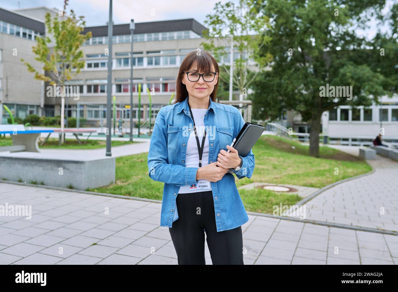 Schullehrerin mittleren Alters mit digitalem Tablet posiert in der Nähe des Schulgebäudes Stockfoto
