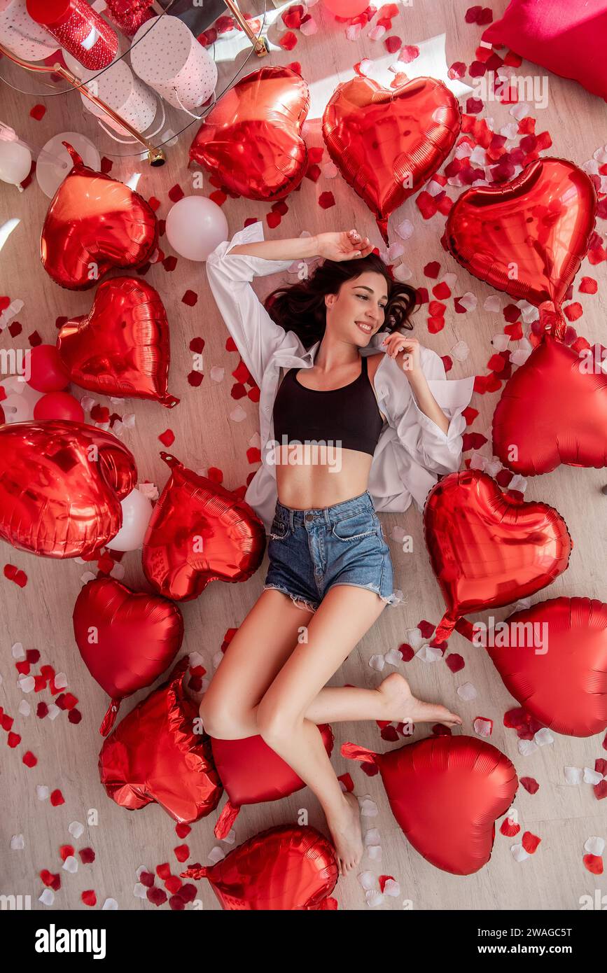Blick von oben auf die junge Frau, die am Valentinstag auf Holzboden liegt und von roten Herzballons umgeben ist. Das Mädchen lächelt und trägt ein lässiges Outfit. Boden Stockfoto