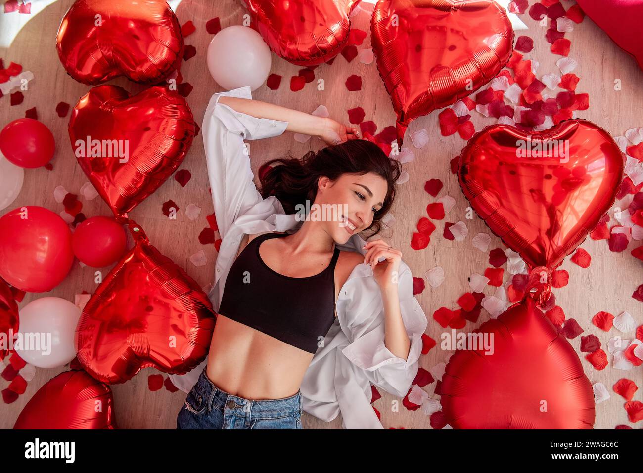 Blick von oben auf die junge Frau, die am Valentinstag auf Holzboden liegt und von roten Herzballons umgeben ist. Das Mädchen lächelt und trägt ein lässiges Outfit. Boden Stockfoto