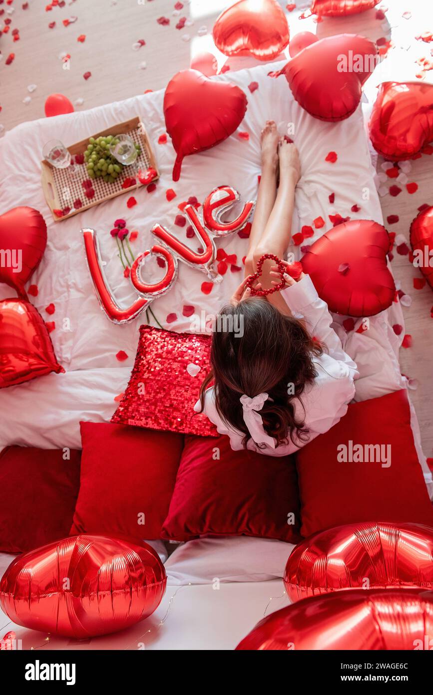 Blick von oben auf eine junge Frau, die um rote Ballons und Rosenblätter auf dem Bett sitzt. Gesichtsloser Blogger hält herzförmige Perlen. Dekoration mit der Aufschrift Love, Stockfoto