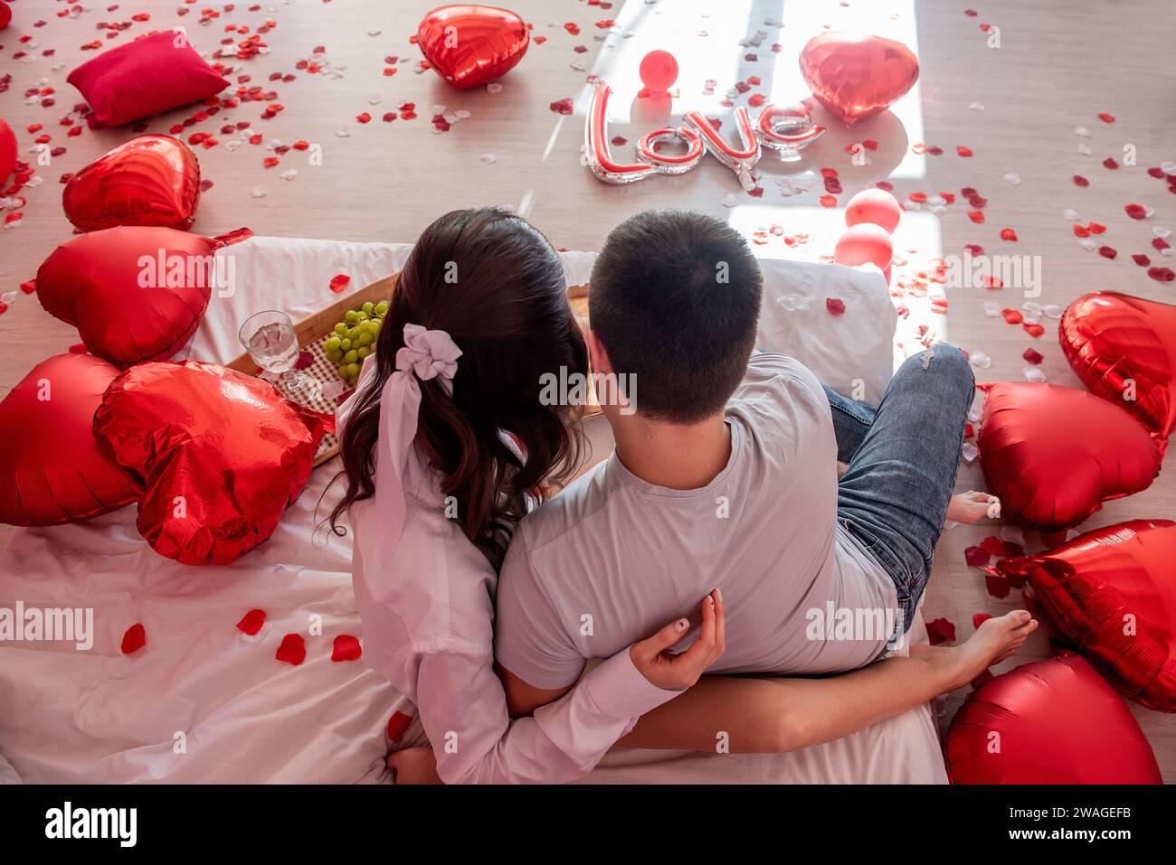 Blick von oben auf junges Paar, das sich mit roten Herzballons auf dem Bett gemütlich und intim fühlt. Mann und Frau umarmen sich gegenseitig in Valenz Stockfoto