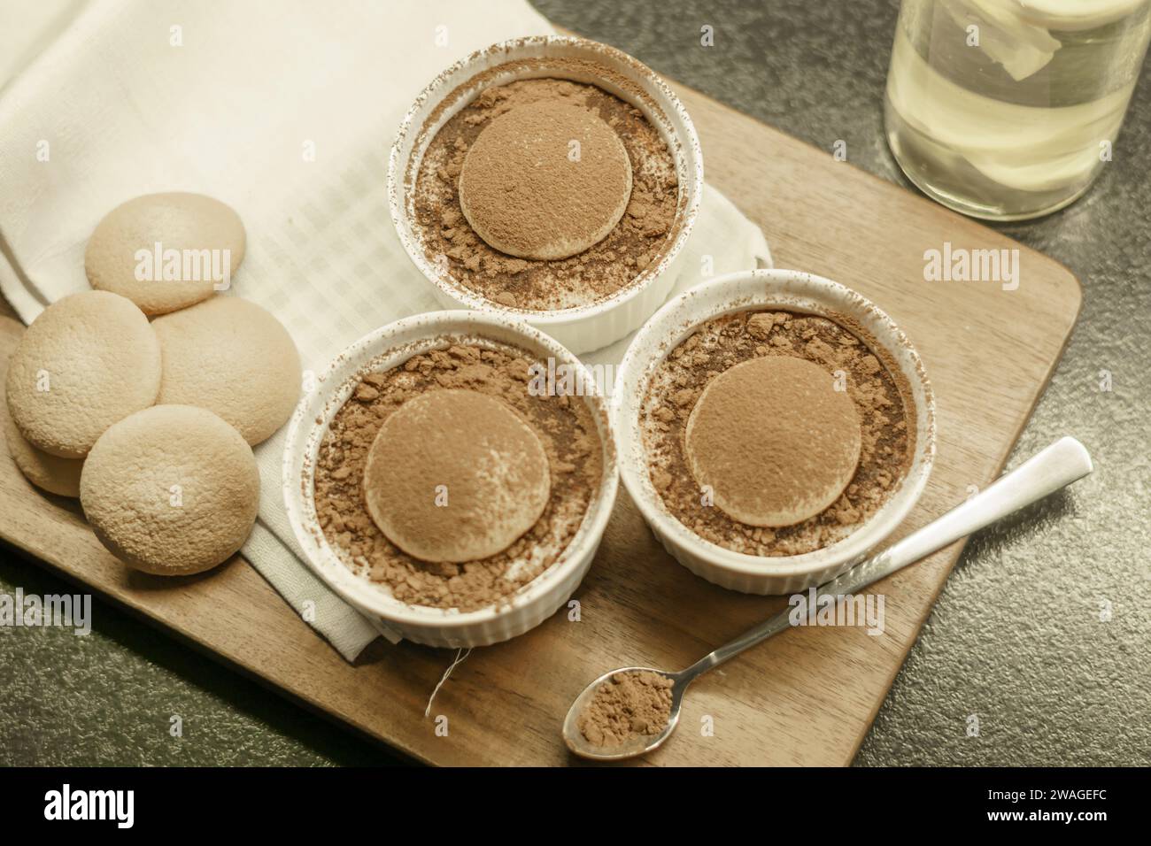 Schüsseln mit hausgemachtem Tiramisu-Dessert auf einem Holzbrett, Löffel mit Kakaopulver, Biskuitkuchen und einem Tuch Stockfoto