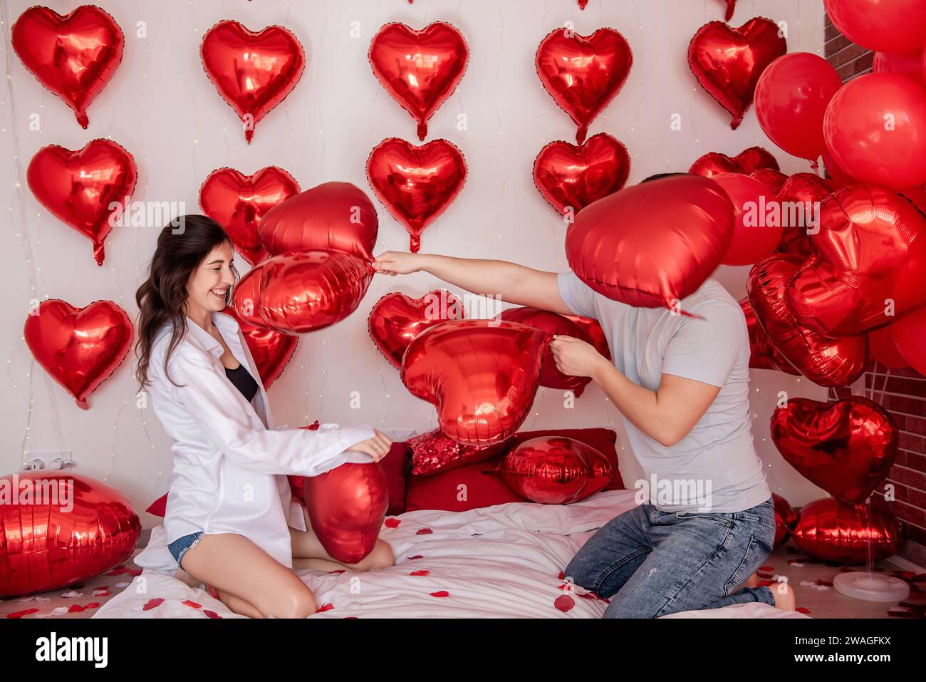 Verspielter Moment zwischen Paaren, die sich in Kissenballons auf dem Bett kämpfen, mit herzförmigen roten Ballons im Hintergrund. Die Frau macht fröhlich rumtäuscht Stockfoto