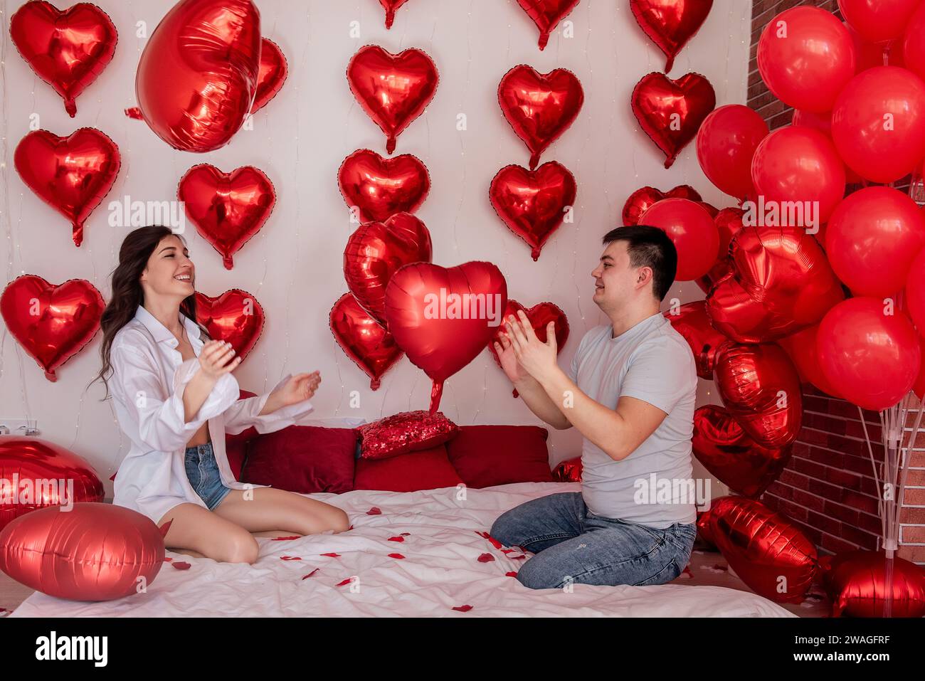 Verspielter Moment zwischen Paaren, die sich in Kissenballons auf dem Bett kämpfen, mit herzförmigen roten Ballons im Hintergrund. Die Frau macht fröhlich rumtäuscht Stockfoto