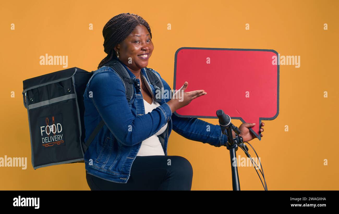 afroamerikanische Lieferfrau mit Rucksack-Bewegung auf rotem Schild in der Hand. Freundlicher Fahrradmessenger mit roter Sprechblase für eine zuverlässige, umweltfreundliche App zur Lebensmittellieferung. Stockfoto