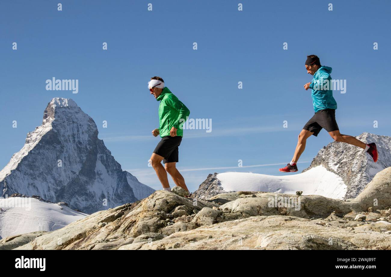 Seitenansicht von zwei Läufern in Sportbekleidung, die auf einem hohen Bergrücken mit schneebedeckten Gipfeln in der Ferne joggen Stockfoto
