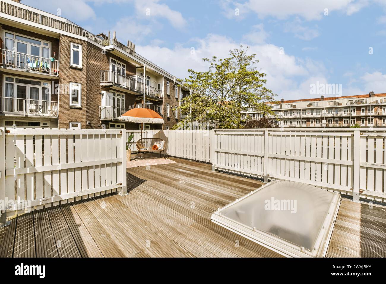 Holzterrasse mit Sofa und orangefarbenem Sonnenschirm und weißem Zaun vor dem Appartementgebäude an sonnigen Tagen Stockfoto