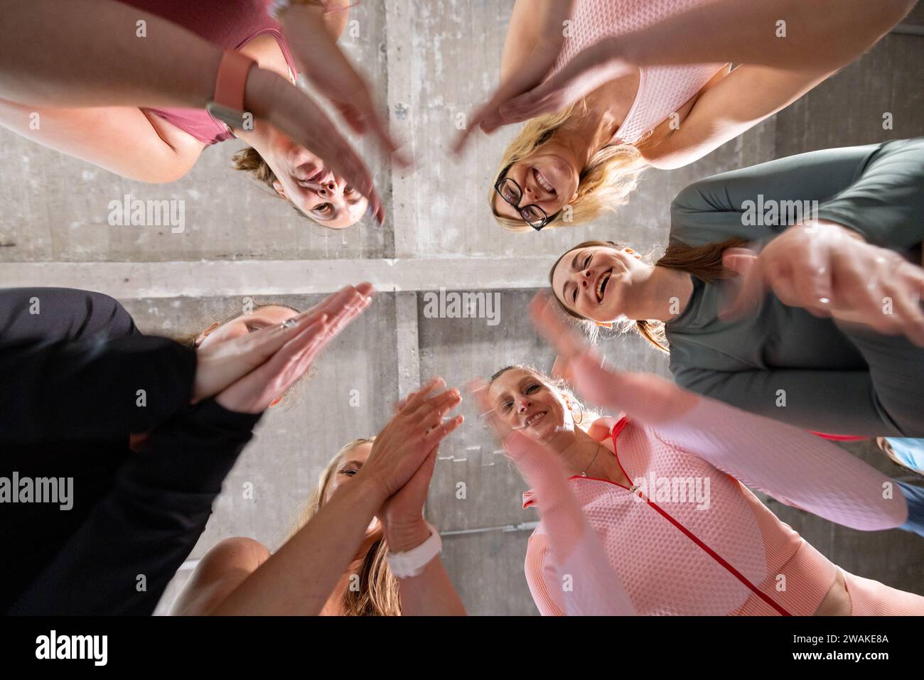 Das Bild bietet eine aufmunternde Perspektive einer Gruppe von Frauen in einer Zusammenkunft, von unten betrachtet. Ihre erhabenen Hände treffen sich in der Mitte, symbolisieren Einheit und kollektive Motivation. Die Frauen lächeln und zeigen Freude und positive Energie in der Gruppe. Sie tragen Sportbekleidung, was darauf hindeutet, dass sie Teil einer Fitness- oder Teambuilding-Aktivität sind. Die darüber liegende Betondecke und die Annäherung ihrer Hände am Brennpunkt unterstreichen die Stärke und die Stütze, die sie voneinander schöpfen. Motivationskreis: Team Huddle von unten. Hochwertige Fotos Stockfoto