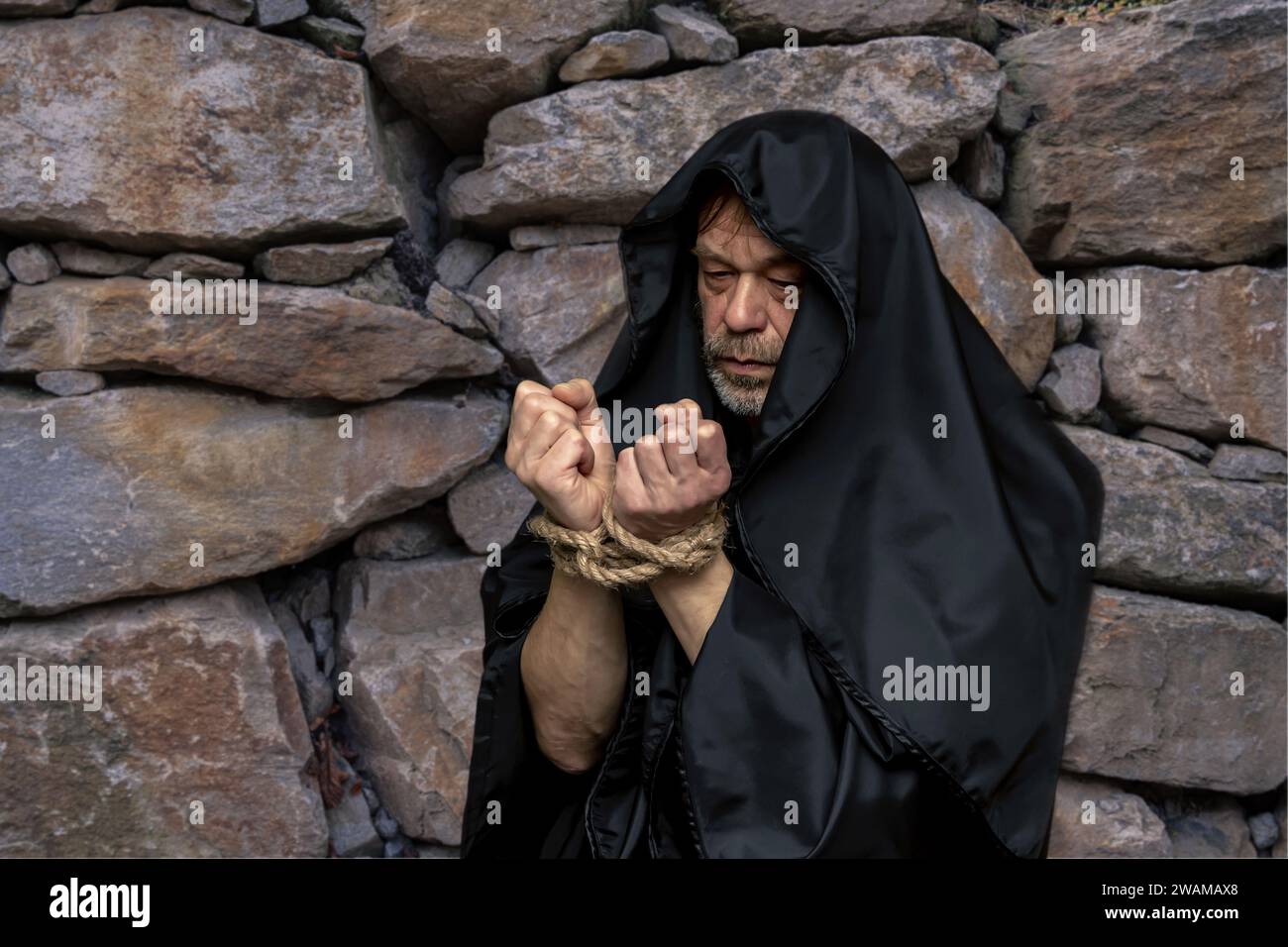 Ein älterer Mönch, dessen Hände mit einem Seil an eine Steinmauer gebunden waren. Stockfoto