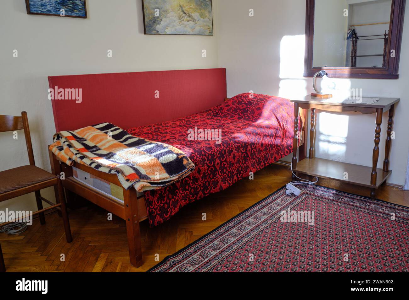 Altes Schlafzimmer mit kleinem Einzelbett, rustikaler roter Bettdecke und Teppich, beleuchtet von hellem Sonnenschein vom Fenster. Stockfoto