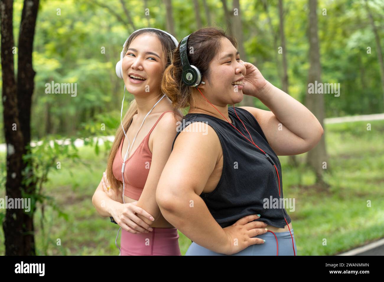 Zwei glückliche asiatische Freundinnen, eine übergroße und eine schlanke, stehen mit dem Rücken zusammen und hören Musik von ihren Kopfhörern in einem lokalen Park Stockfoto