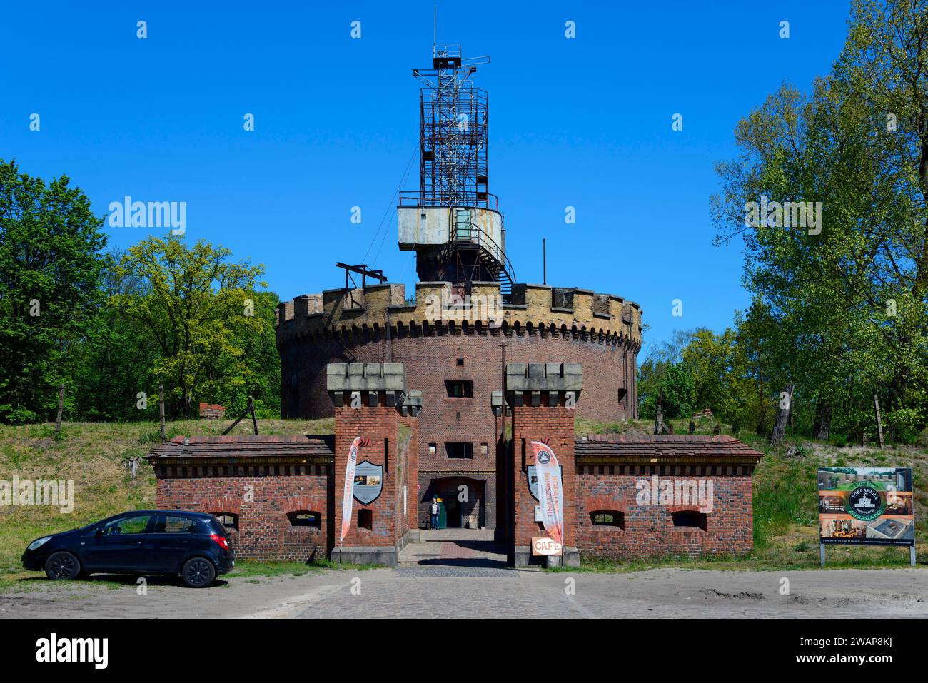 Alter Backsteinturm mit Antenne unter klarem blauem Himmel, ein Auto parkt, umgeben von Bäumen im Frühling, Castel Sant'Angelo, Fort Aniola, Swinoujscie, Swinoujs Stockfoto