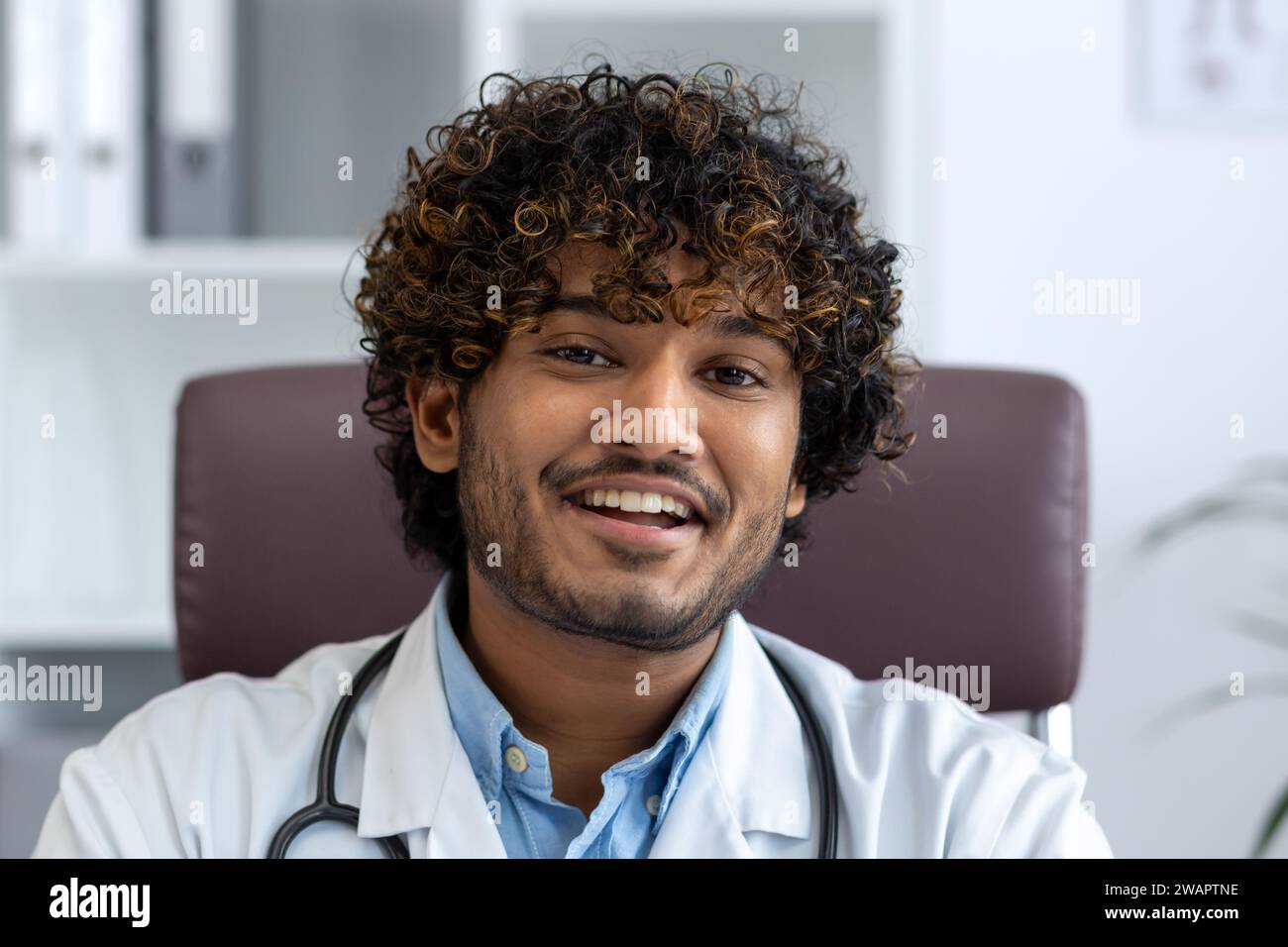 Close-up Porträt des Arztes in der Klinik am Arbeitsplatz, Mann in weißem medizinischen Mantel lächelnd in die Kamera, zufriedener indischer Mann mit lockigen Haaren. Stockfoto