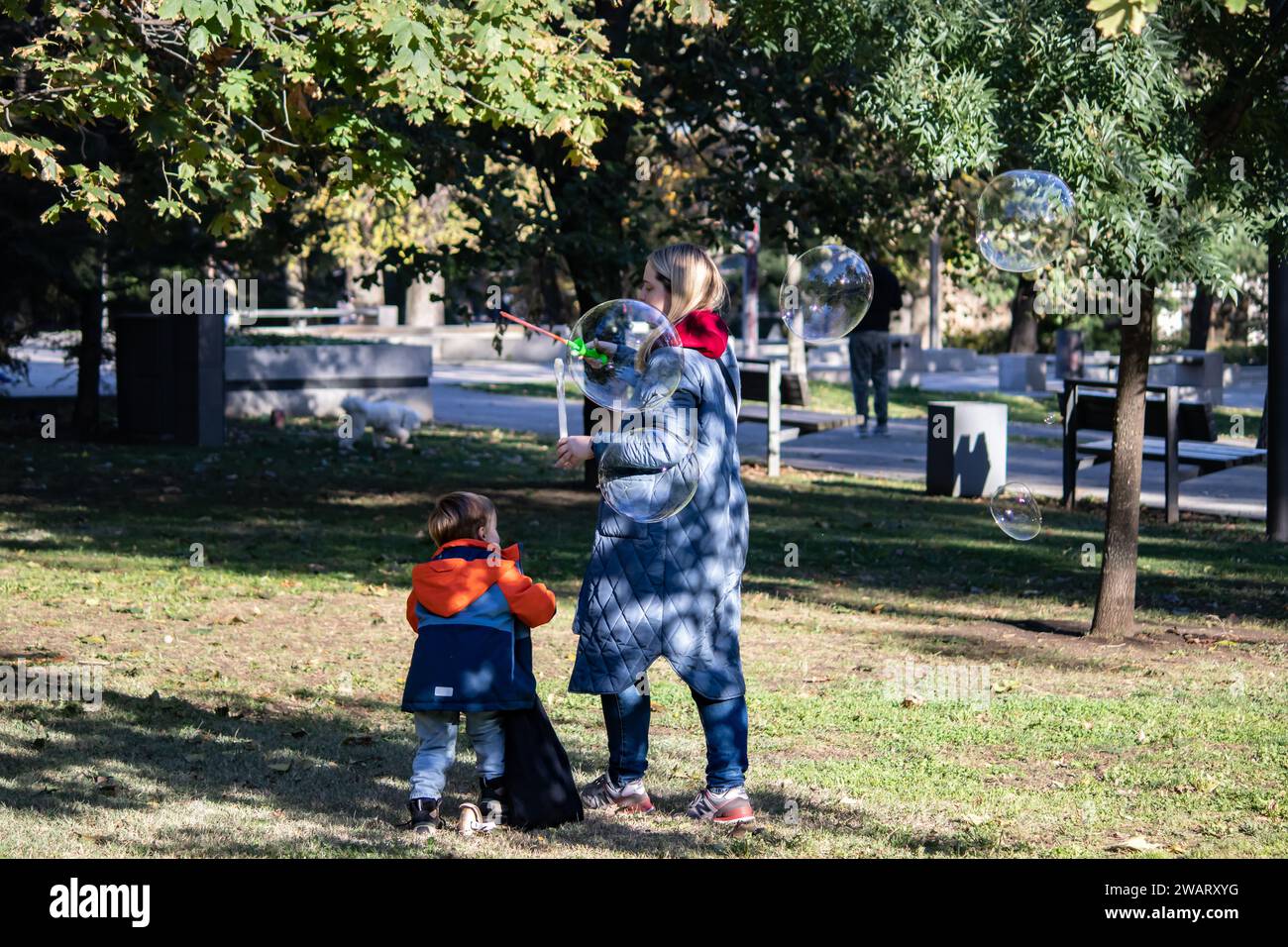 Mutter mit ihrer kleinen Sonne spielt im öffentlichen Stadtpark, macht Seifenballons, fröhlicher Junge rennt herum Stockfoto