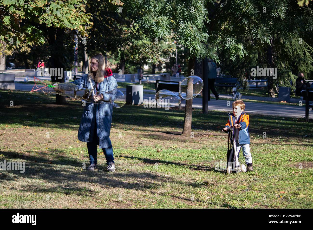 Mutter mit ihrer kleinen Sonne spielt im öffentlichen Stadtpark, macht Seifenballons, fröhlicher Junge rennt herum Stockfoto