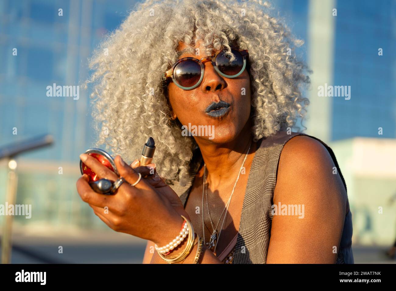 Ausdrucksstarke, schwarze Dame mit grauem Afro-Haar und Sonnenbrille auf blauem Lippenstift im sonnigen Stadtzentrum. Konzept: pro-Aging, Selbstvertrauen Stockfoto