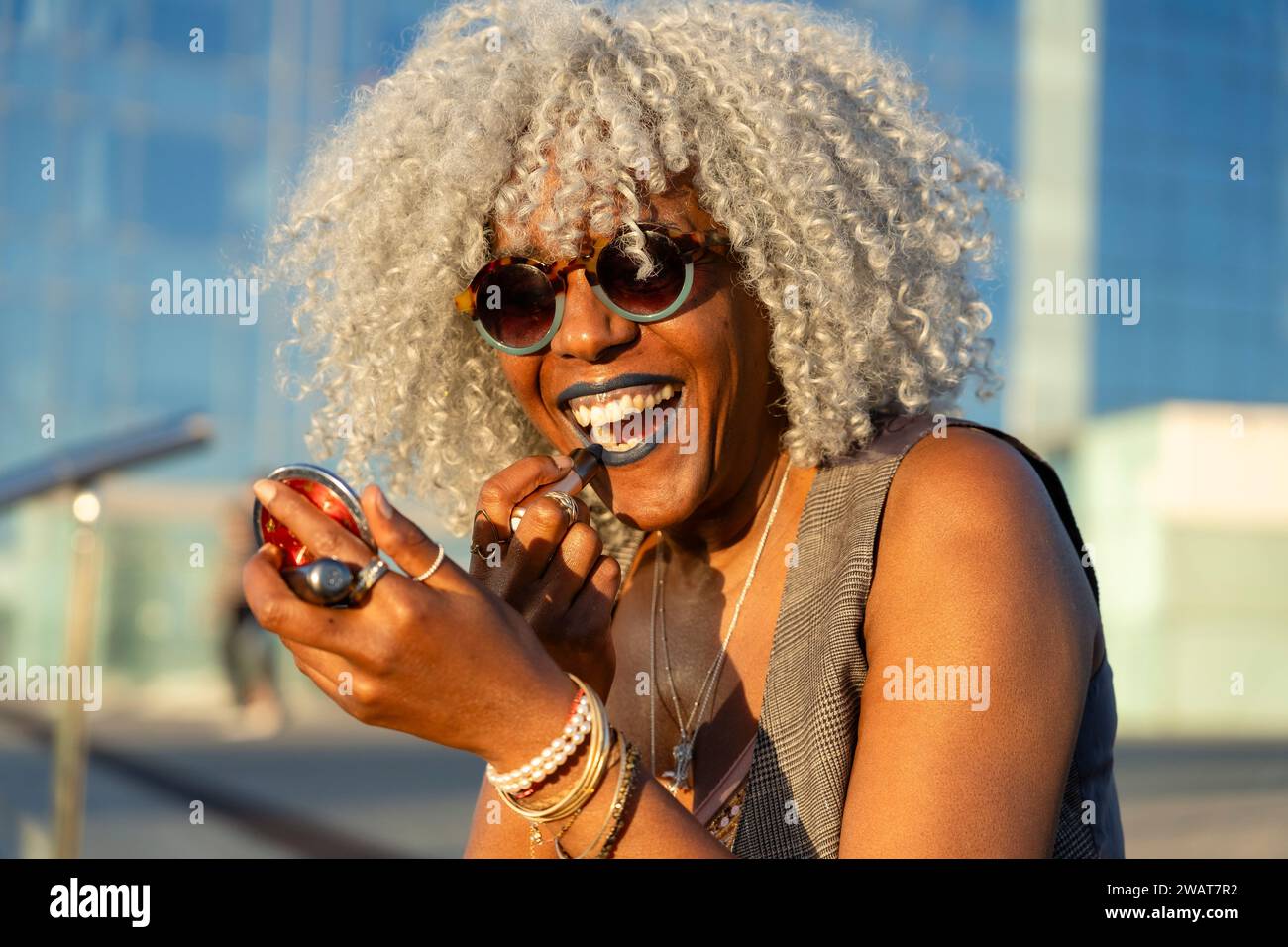 Ausdrucksstarke, schwarze Dame mit grauem Afro-Haar und Sonnenbrille auf blauem Lippenstift im sonnigen Stadtzentrum. Konzept: pro-Aging, Selbstvertrauen Stockfoto