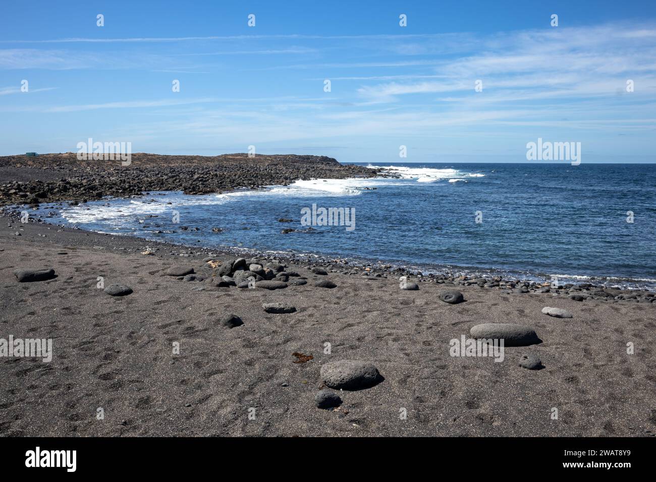 Strand mit dunklem Sand, aus Lava. Große Kieselsteine. Ruhiges Wasser des Atlantischen Ozeans. Blauer Himmel mit hellweißen Wolken. Lanzarote, Kanarische Inseln, Stockfoto