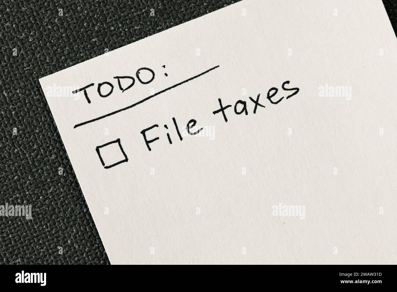 Handgeschriebene To-Do-Liste mit „File Taxes“-Erinnerung und einem Kontrollkästchen auf einer weißen Notiz vor einem grauen texturierten Hintergrund, flache Ansicht von oben nach unten Stockfoto