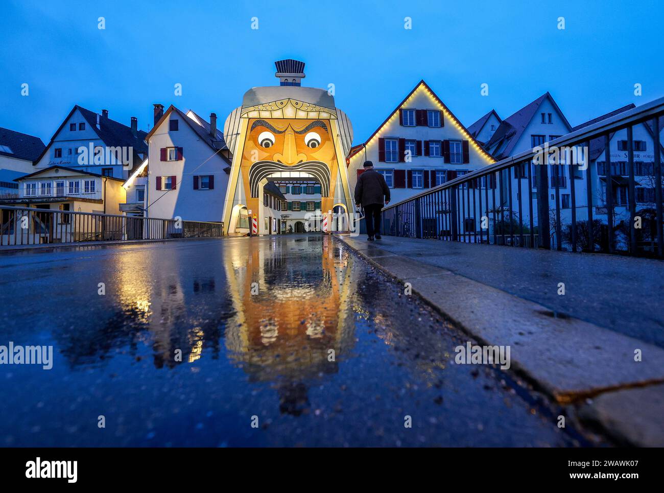 07. Januar 2024, Baden-Württemberg, Riedlingen: Der Goletor steht am Eingang zur Altstadt von Riedlingen und spiegelt sich in einer Regenpfütze wider. Das Tor zeigt den überdimensionalen Kopf der „Gole“, eine der Riedlinger Fasnet-Figuren aus dem Jahr 1890. Das Tor wird nur zu besonderen Anlässen in der Donaustadt errichtet. Grund dafür ist die Karnevalssitzung der Vereinigung Schwäbisch Alemannischer Narrenzünfte (VSAN) am 04.02.2024. Wer in die Riedlinger Innenstadt kommt, sieht nun die rollenden Augen der mächtigen „Gole“ aus der Ferne und kann nur noch die historische Altstadt betreten Stockfoto
