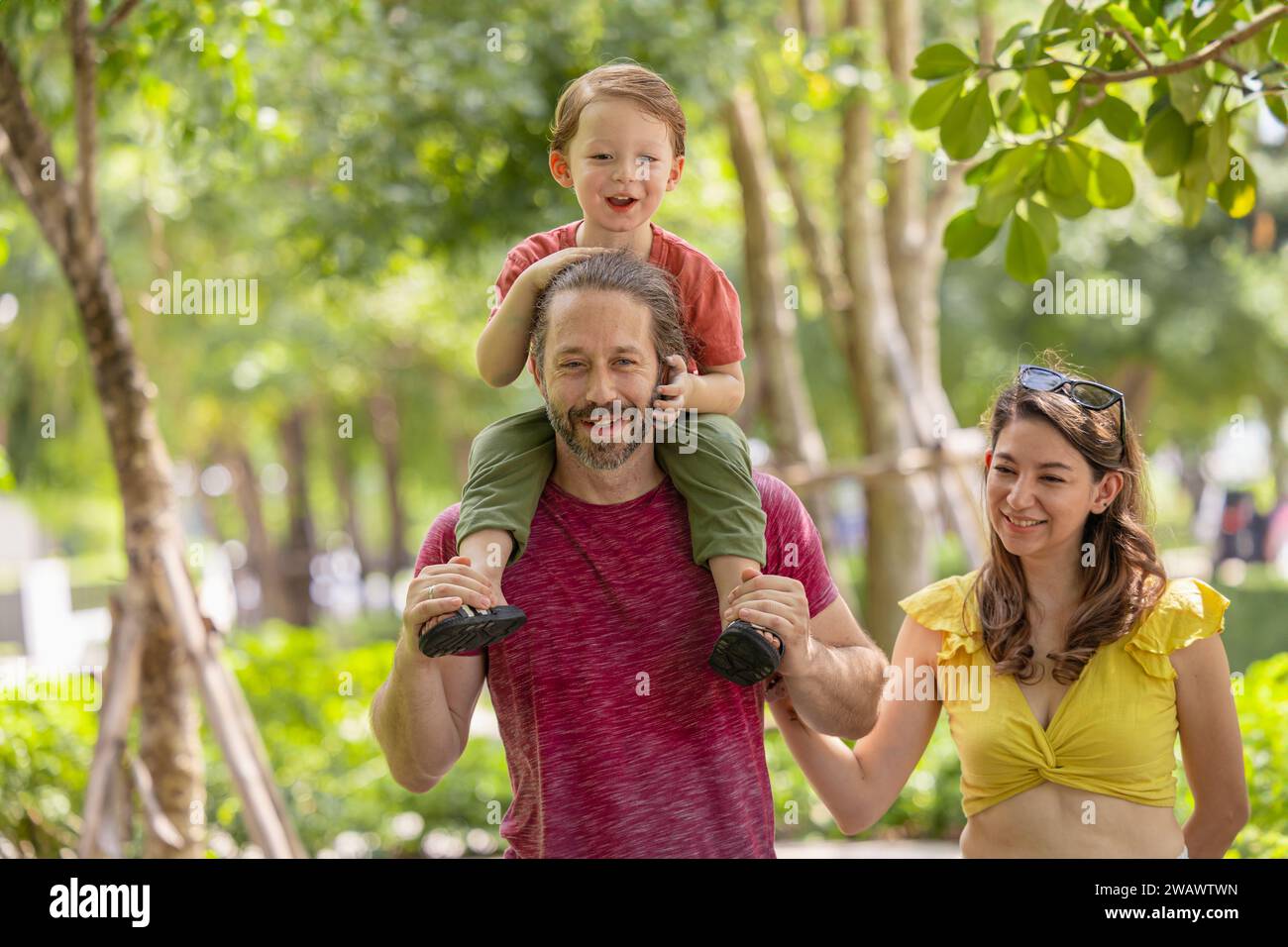 Glückliche Familie zusammen spazieren im öffentlichen grünen Park junger Junge, der auf der Schulter des Vaters um frische ökologisches Bäume herum sitzt. Stockfoto