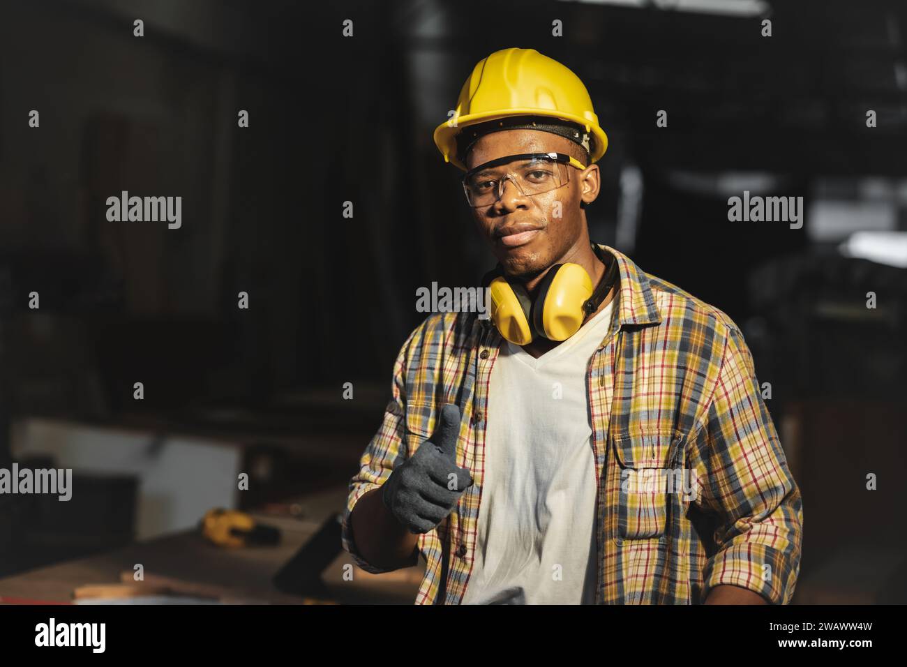 afrikanischer schwarzer Arbeiter in der Werkstatt mit Schutzhelm und Ohrenschützer stehend Daumen nach oben schauend Kamera Stockfoto