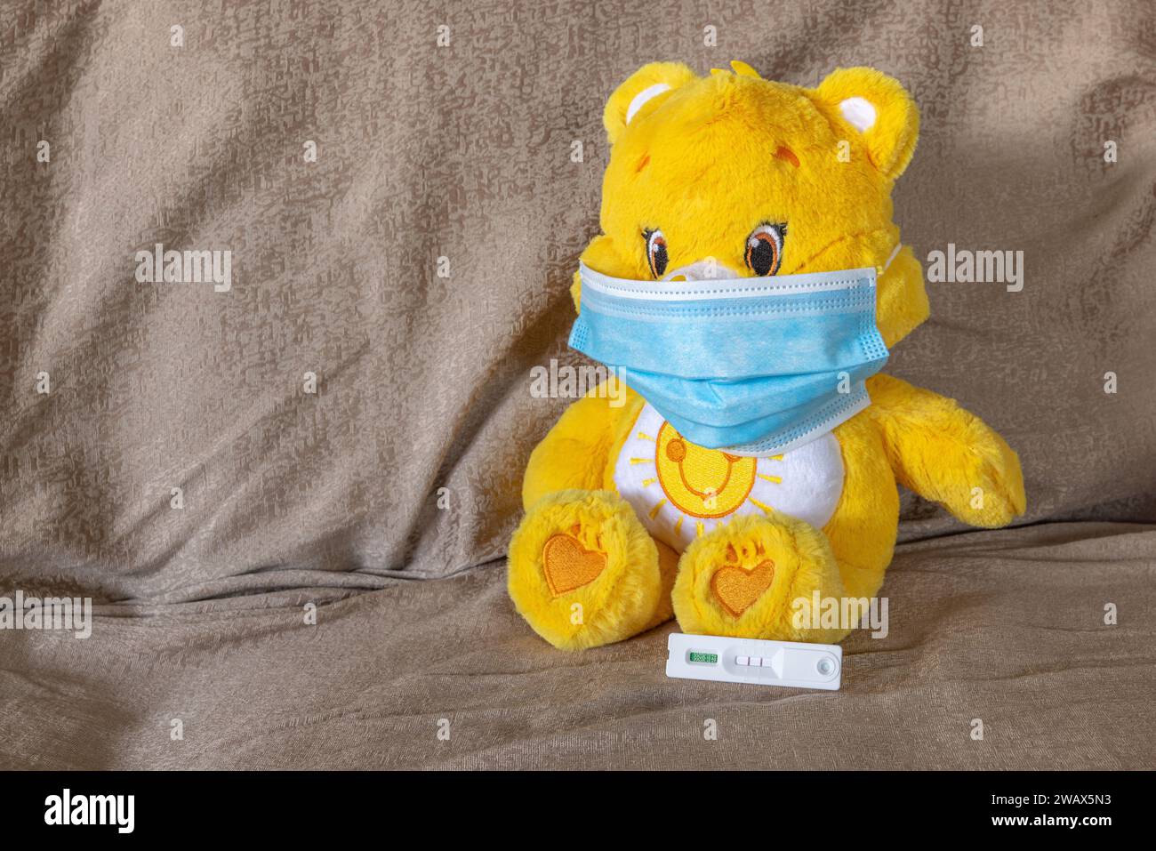 Nahaufnahme eines gelb-weiß gefärbten Teddybären auf einem Sofa mit blauer Gesichtsmaske während der COVID-19-Pandemie mit positivem Antigenschnelltest. Stockfoto