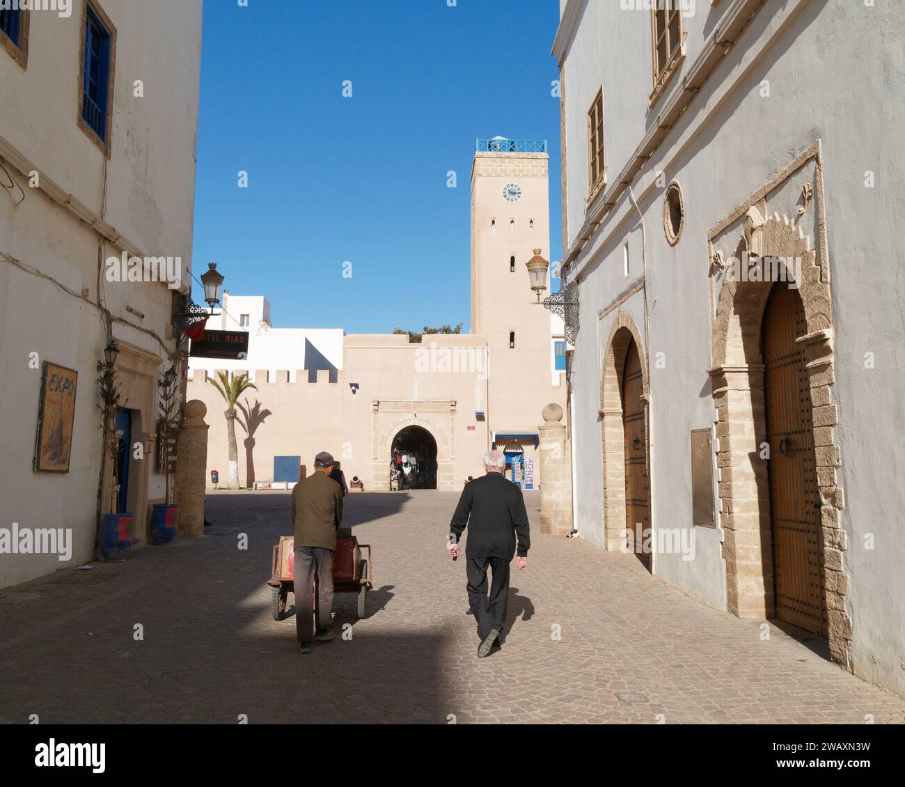 Der Mann schiebt einen Wagen in einer Straße in der historischen Medina mit dem Uhrenturm vor der Stadt Essaouira, Marokko. Januar 2024 Stockfoto