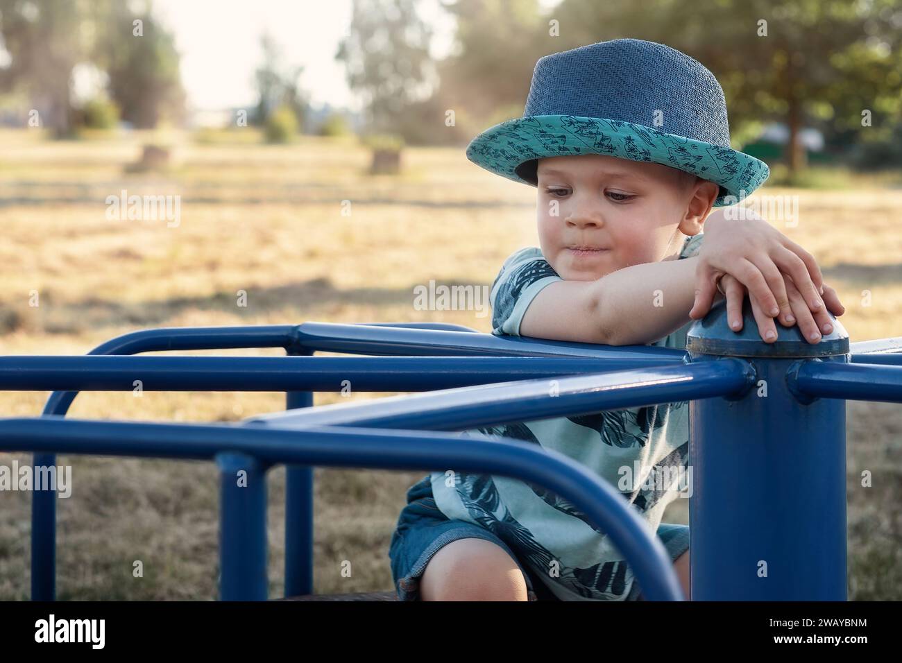 Ein ernster und nachdenklicher kleiner Junge posiert für einen Fotografen an einem sonnigen Tag in einem blauen Outdoor-Karussell. Stockfoto