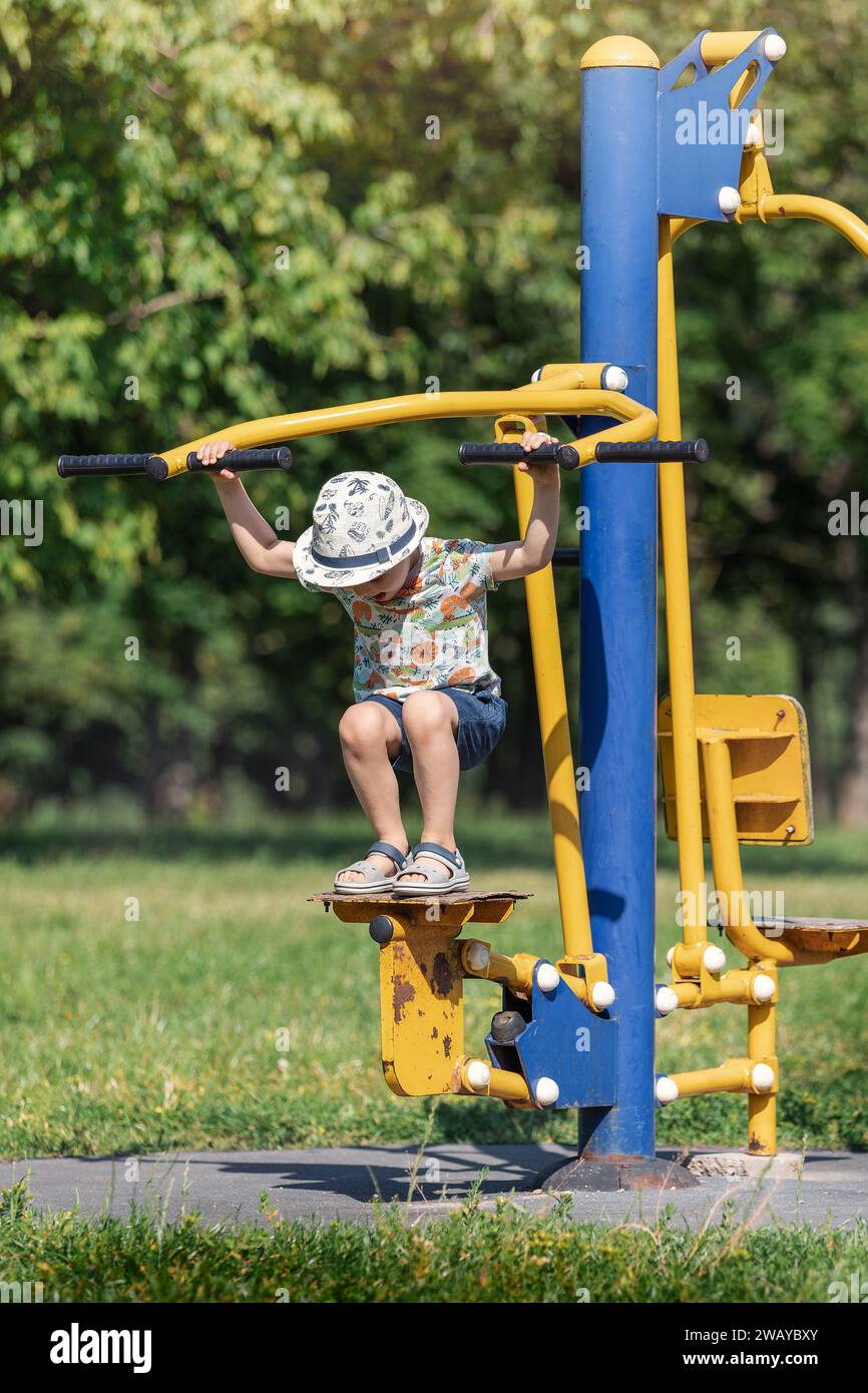 Ein kleiner Junge mit Hut trainiert im Park auf einem gelb-blauen Metall-Outdoor-Trainingsgerät. Stockfoto