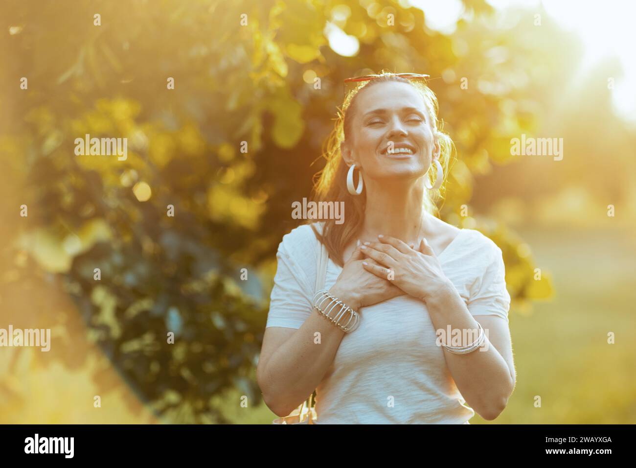 Sommerzeit. Glückliche moderne Frau in weißem Hemd mit Hand auf der Brust draußen. Stockfoto