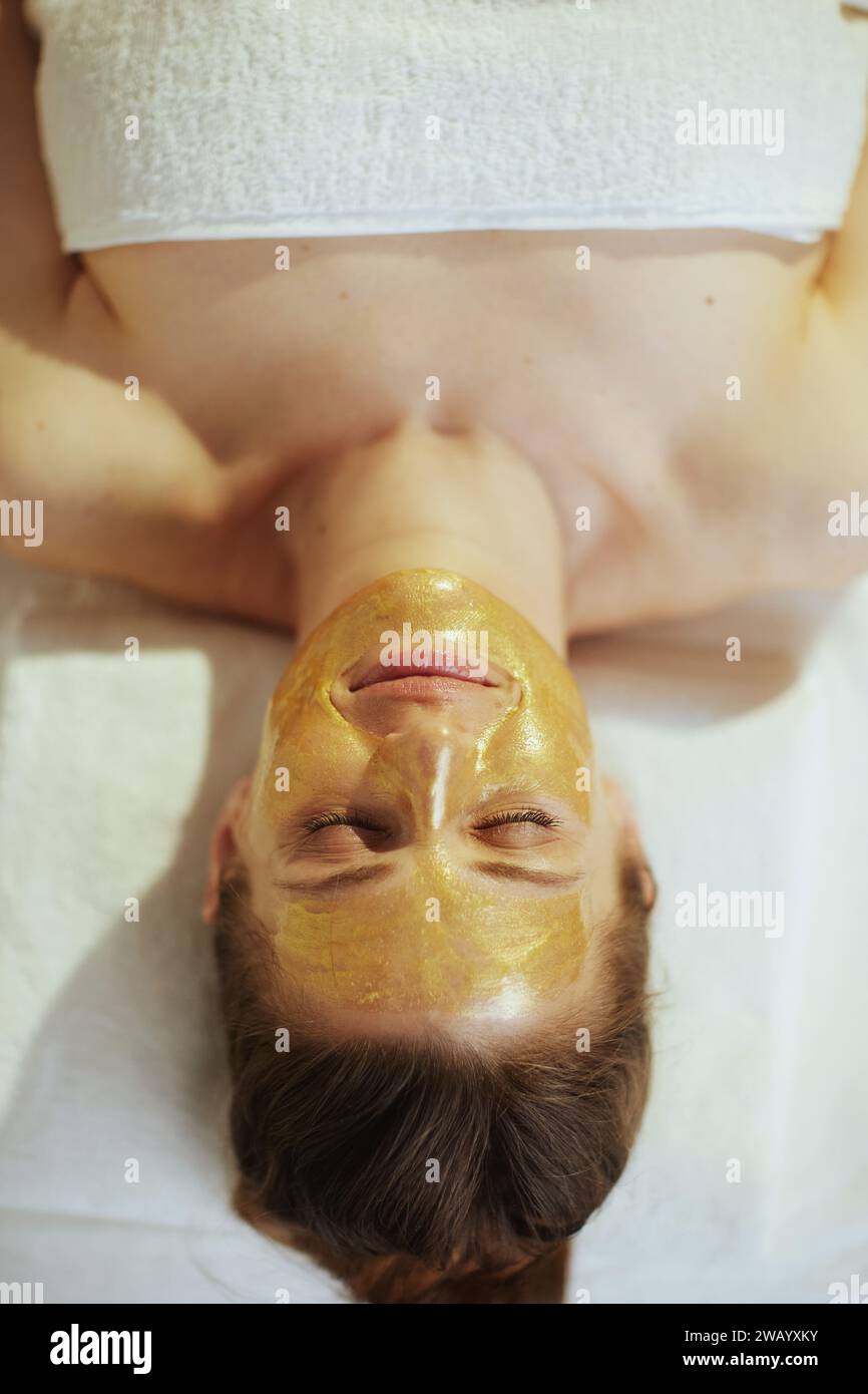 Gesundheitszeit. Obere Ansicht einer entspannten modernen Frau in Massagekabine mit goldener Kosmetikmaske auf dem Gesicht, das auf Massagetisch liegt. Stockfoto