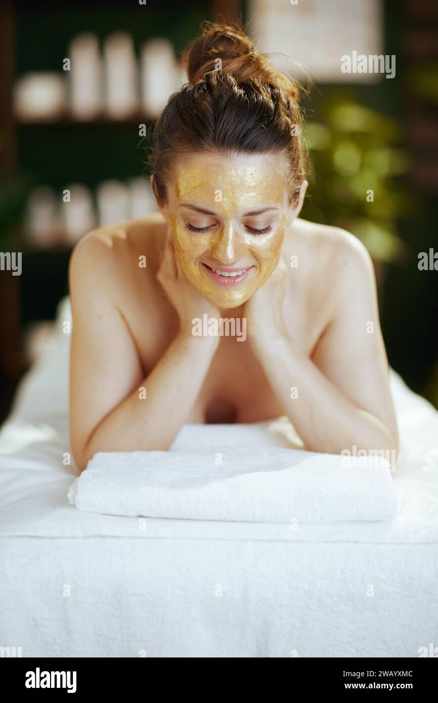 Gesundheitszeit. Glückliche, moderne Frau im Spa-Salon mit goldener Kosmetikmaske auf dem Gesicht, die auf dem Massagetisch liegt. Stockfoto