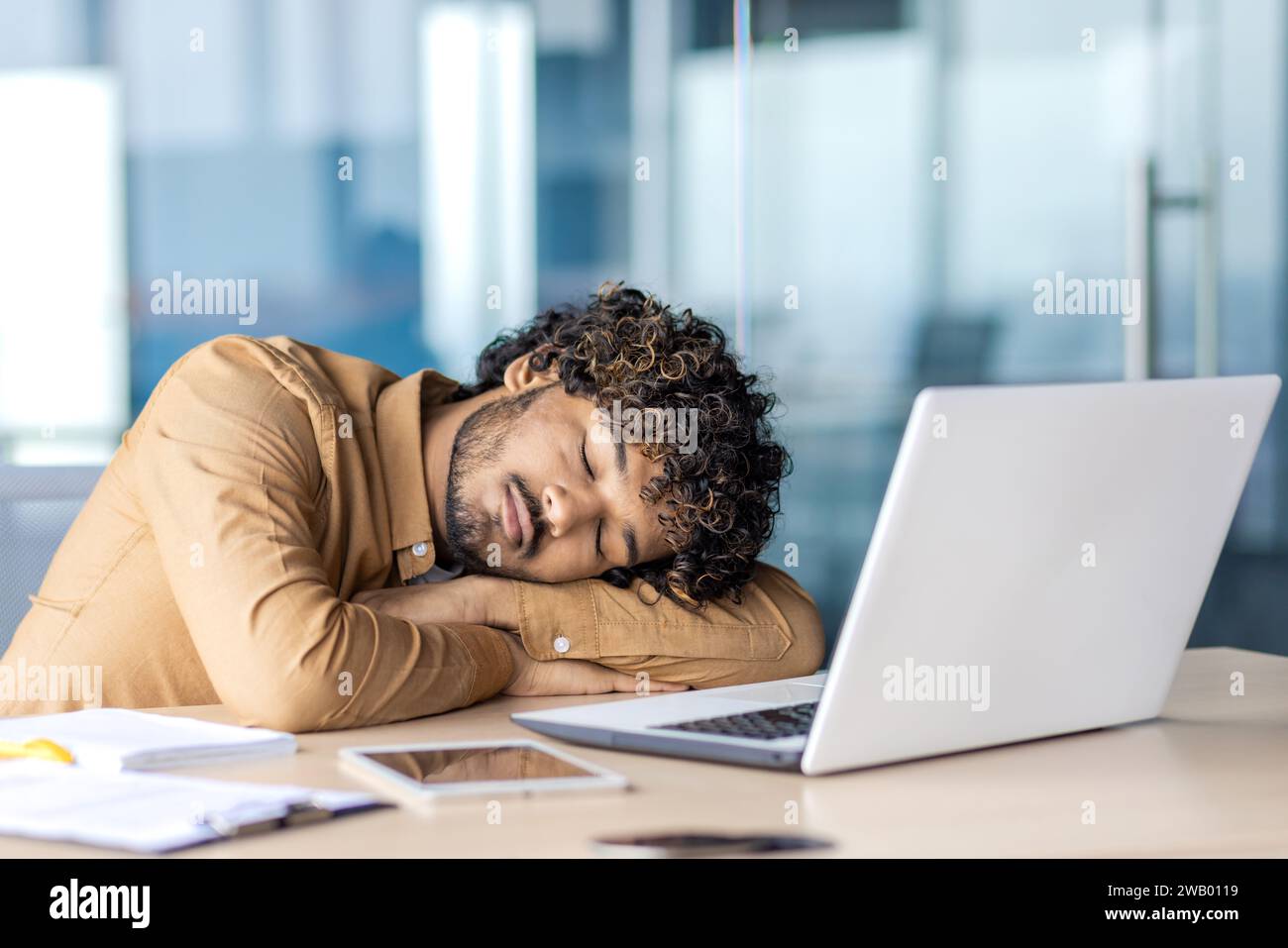 Geschäftsmann, der am Arbeitsplatz schläft, junger übermüdeter und überarbeiteter Mann, der auf dem Schreibtisch im Büro mit Laptop liegt, lange Arbeit bis spät, Abgabetermin am Abend. Stockfoto
