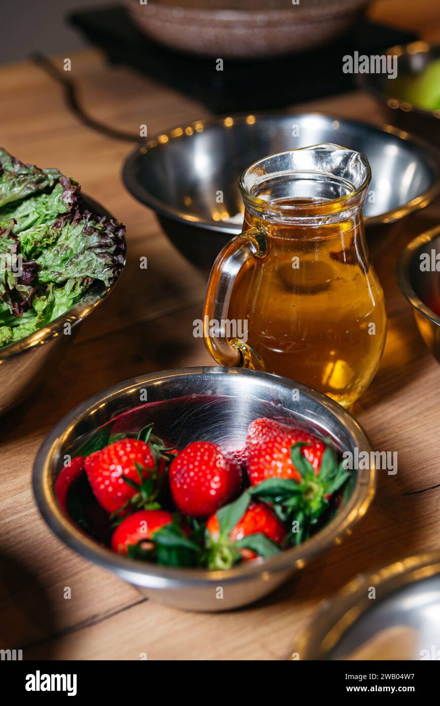 Eine Schüssel aus Edelstahl mit Reifen Erdbeeren und einem frischen grünen Salat gepaart mit einer Kanne Öl, auf einer hölzernen Küchenzeile angeordnet. Stockfoto