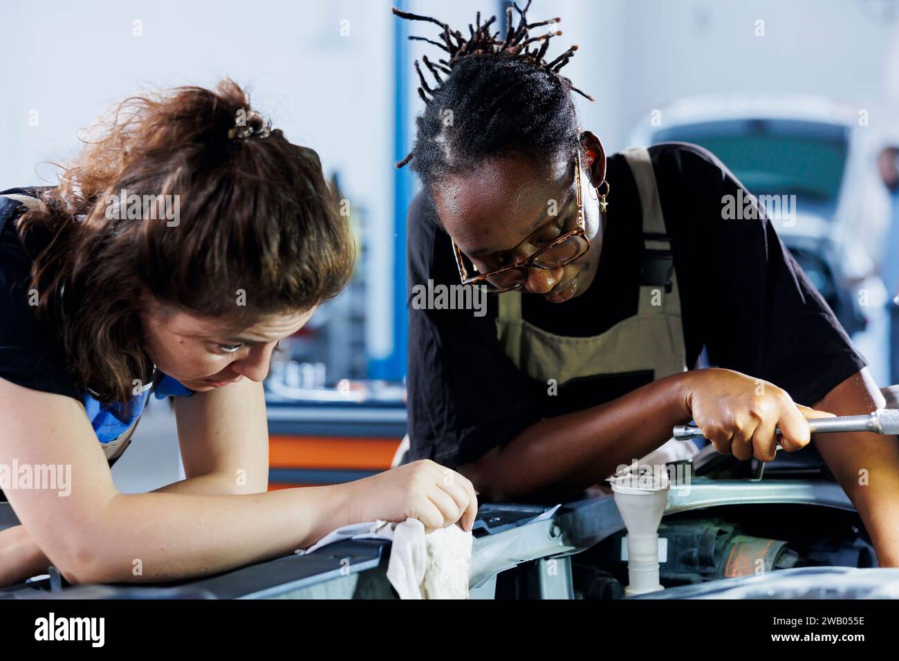 BIPOC-Mechaniker arbeiten an der Wartung defekter Fahrzeuge zusammen und prüfen auf defekten Kühler. Frauen in der Autowerkstatt arbeiten zusammen an der Reparatur des Autos und besprechen die besten Optionen Stockfoto