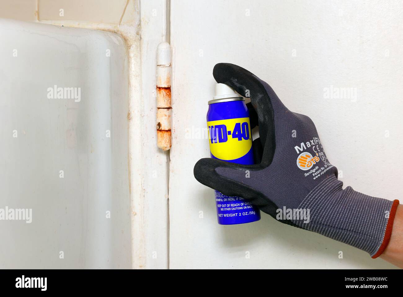 Eine Dose Schmieröl-Spray WD 40 wird von einer Hand mit Handschuhen in der Nähe eines rostigen Türscharniers gehalten. Stockfoto