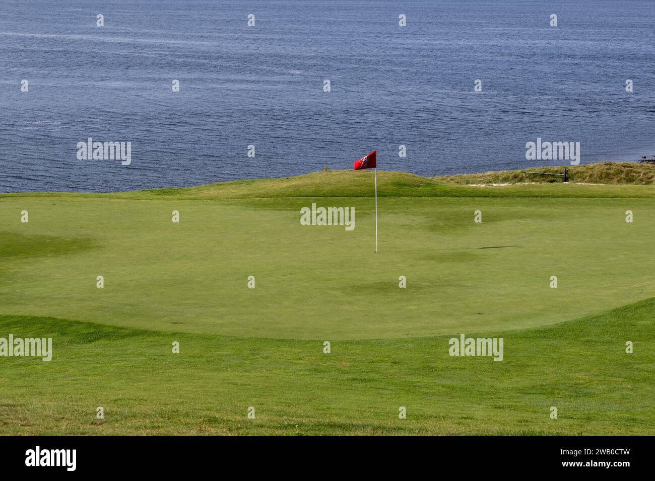 Eine rote und schwarze Flagge auf einem weißen Stock in einem Golfloch am Wasser. Das üppig grüne Gras wird eingetaucht und mit seinem tiefsten Punkt in der Mitte gezüchtet. Stockfoto