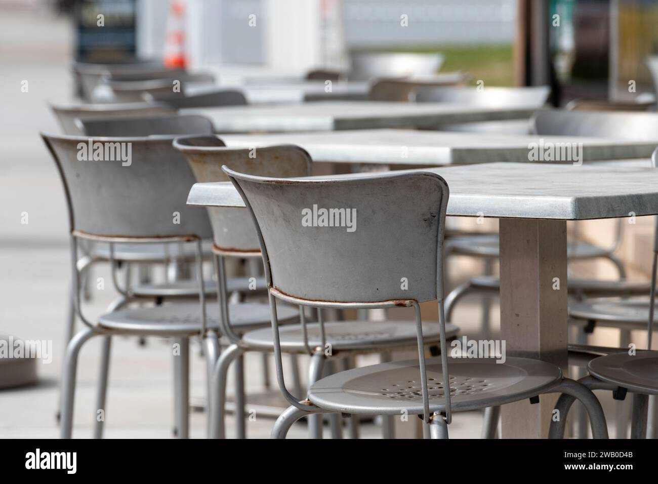 Eine Reihe von leeren quadratischen Tischen und Stühlen aus grauem Metall auf dem Bürgersteig eines Cafés oder einer Bäckerei. Die Stühle haben runde Rücken und sind leer. Th Stockfoto