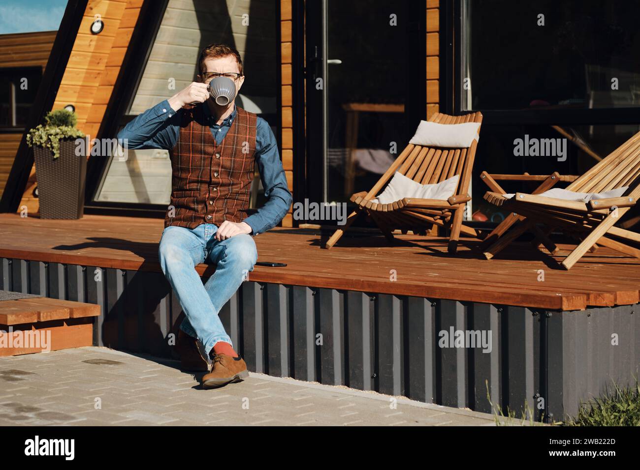 Mann mittleren Alters, der auf der Terrasse einer Holzhütte sitzt und morgens Tee trinkt Stockfoto