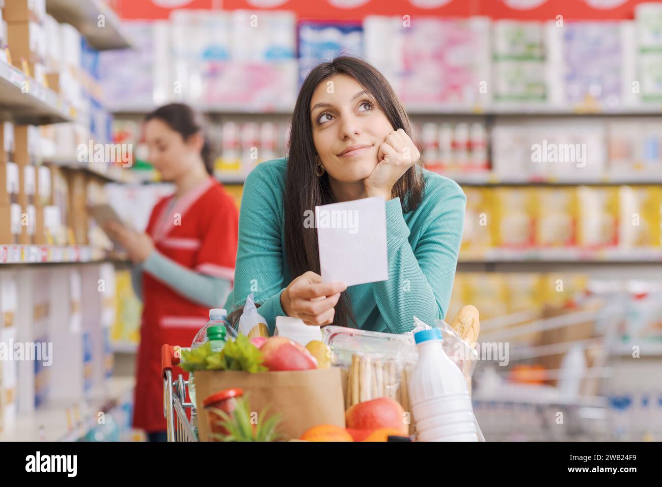Die junge lächelnde Frau, die im Supermarkt einkaufen geht, lehnt sich auf den Wagen, hält eine Lebensmittelliste und lächelt Stockfoto