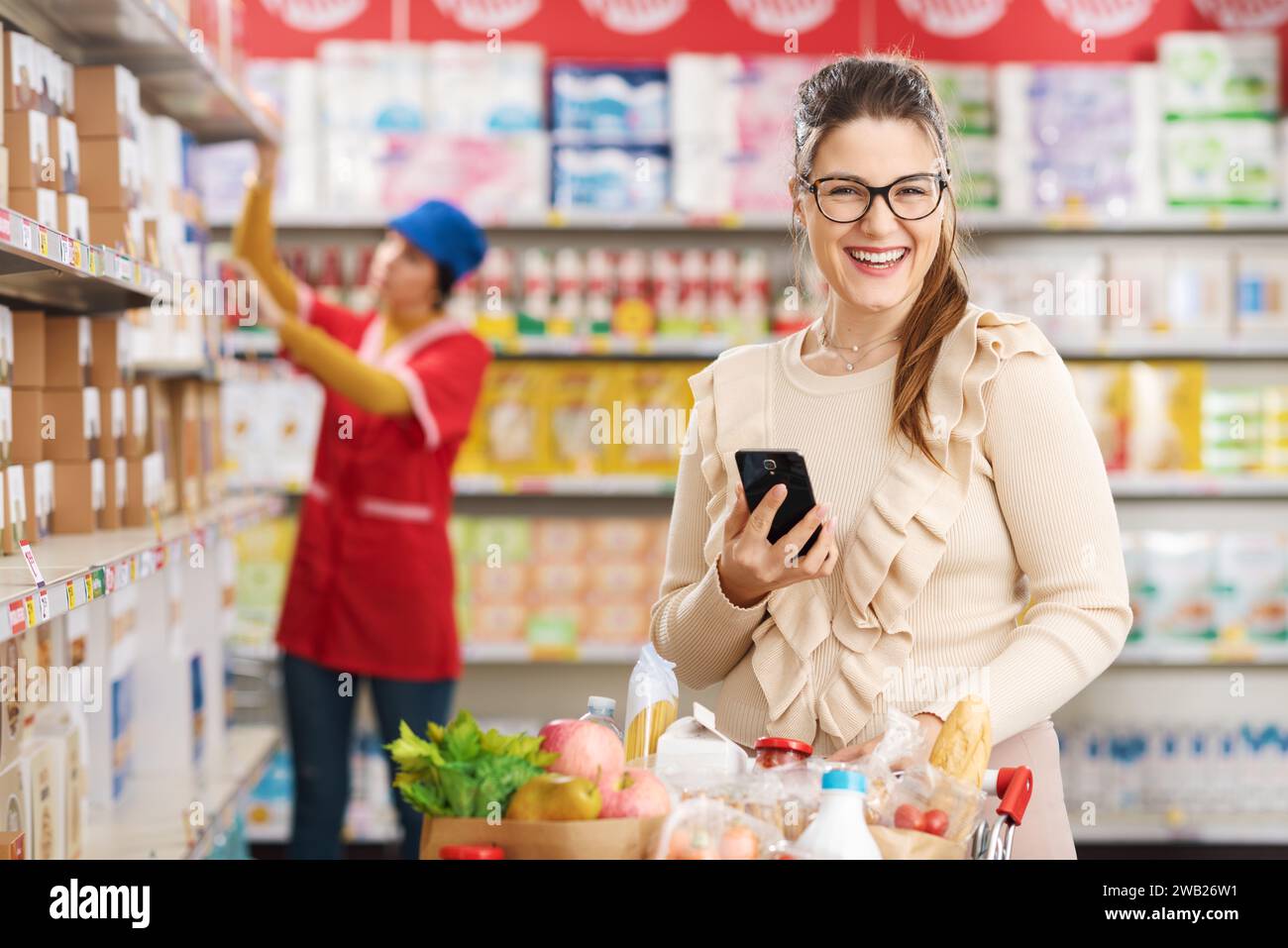 Lächelnde Frau, die Lebensmittel kauft und ein Smartphone im Supermarkt benutzt Stockfoto