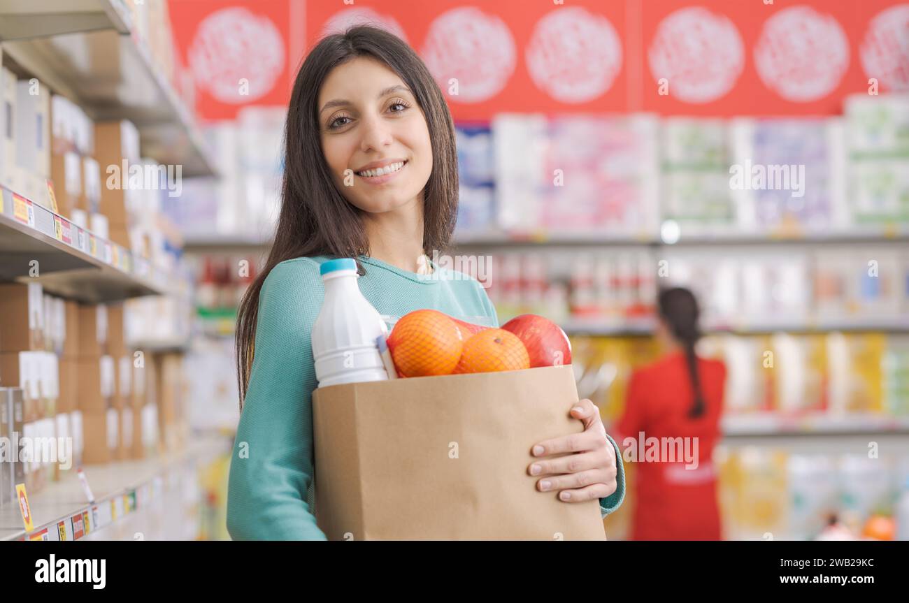 Lächelnde junge Frau, die eine Tasche voller Lebensmittel hält und eine Kamera sieht, eine Verkäuferin arbeitet und Supermarktregale im Hintergrund Stockfoto