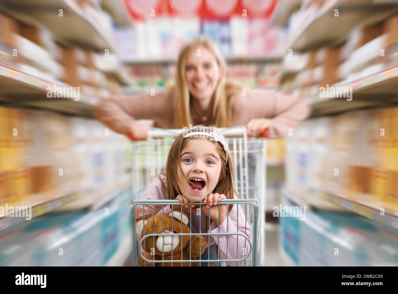 Fröhliches, süßes Mädchen, das im Supermarkt in einem Einkaufswagen sitzt, ihre Mutter schiebt sie: Lebensmittelgeschäft und Lifestyle-Konzept Stockfoto