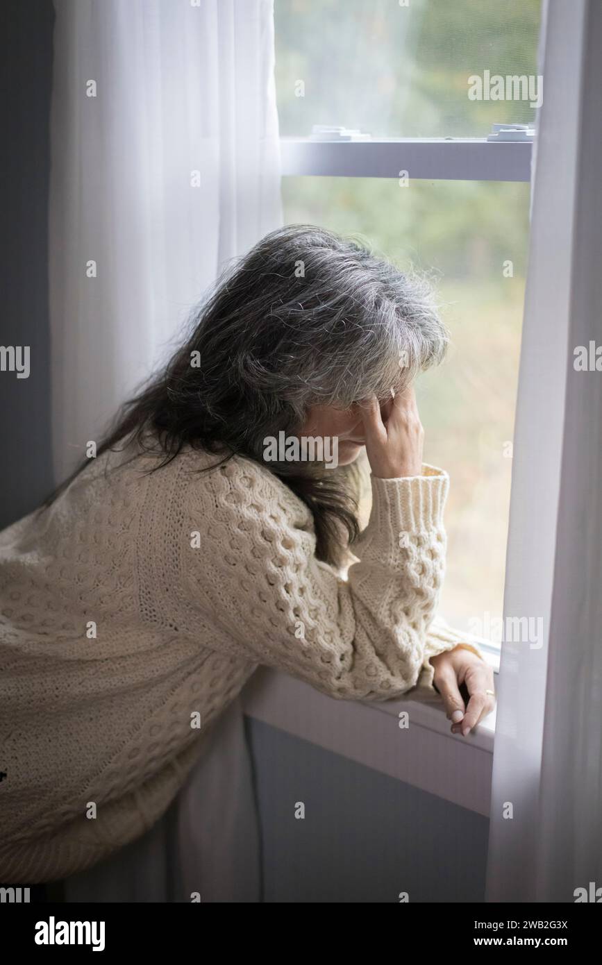 Ältere weiße Frau am Fenster mit Händen über dem Gesicht, die deprimiert aussehen Stockfoto