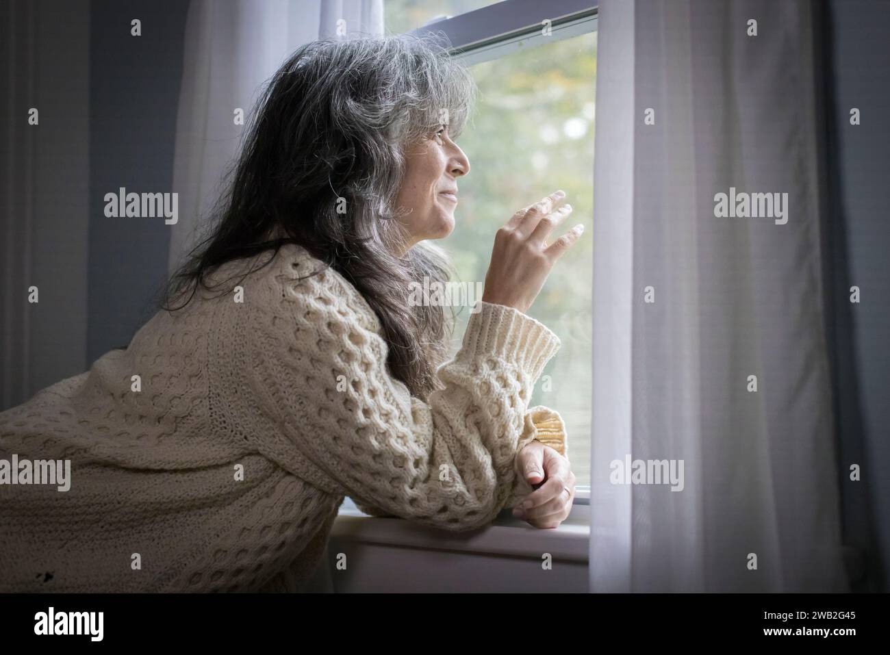 Ältere weiße Frau, die aus dem Fenster blickt, mit fröhlichem Ausdruck Stockfoto