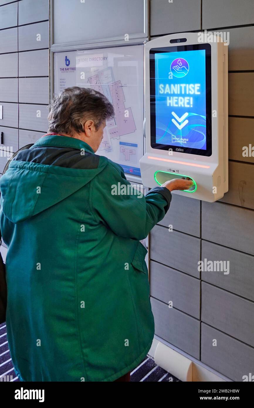 Ältere reife Frau Rückansicht steckt eine Hand in Schlitz an der Wand montierte automatische Desinfektionsgel Dispenser Bildschirm zeigt Werbung Einkaufszentrum Essex England Großbritannien Stockfoto