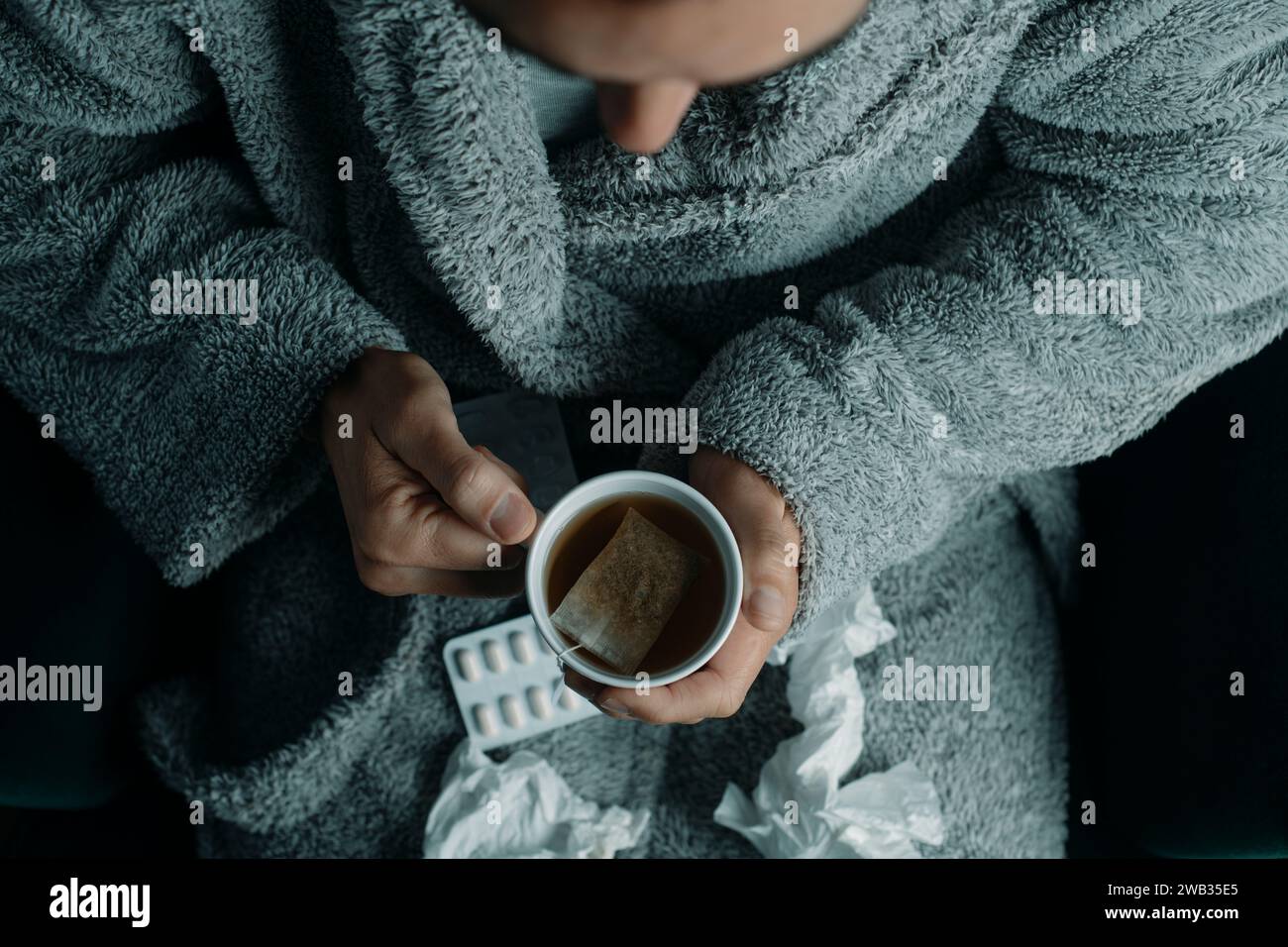 Hochwinkelansicht eines Kranken in einem grauen Hausmantel, zu Hause, sich mit einer Tasse heißem Kräutertee aufwärmen Stockfoto