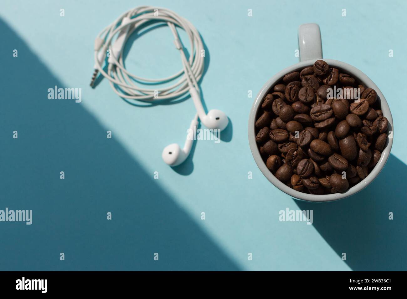 Schwarze Kaffeebohnen in blauer Tasse und weiße Kopfhörer auf blauem Hintergrund mit Blick von oben im Sonnenlicht. Konzept für ein Getränk am Morgen. Kaffee und Musik. Stockfoto